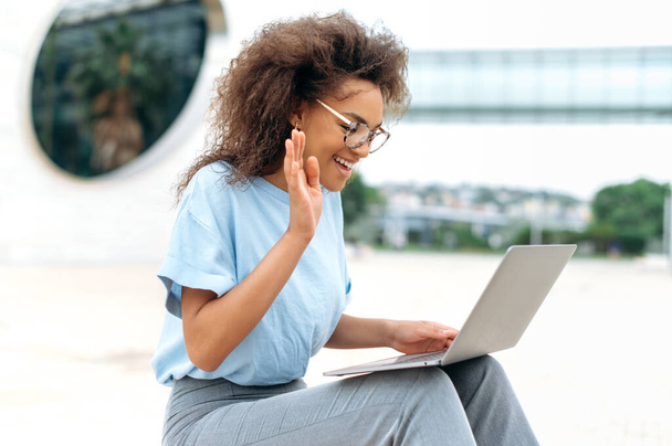 Virtual meeting concept. Busy successful african american young woman, business lady, sitting outdoors, holds online video conference by laptop, discuss financial strategy with colleagues, smiling - Foto, afbeelding