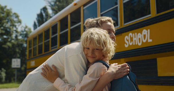 若い笑顔のお母さんは黄色のスクールバスの近くに立って小さな息子を抱いている。明るい朝に搭乗する前に、陽気な母親に別れを告げるバックパックを持つ楽しい巻き少年。女子高生に手を振る幸せな女性. - 写真・画像