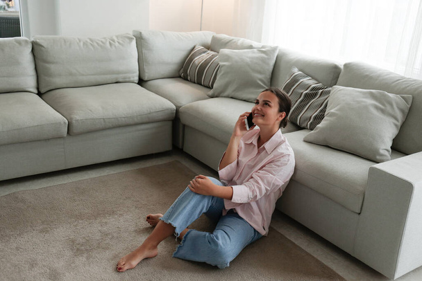Portrait of young brunette woman wearing mom jeans sitting on the floor leaning on the couch talking on the phone and smiling. Joyful female having fun call. Background, copy space, close up. - Foto, immagini