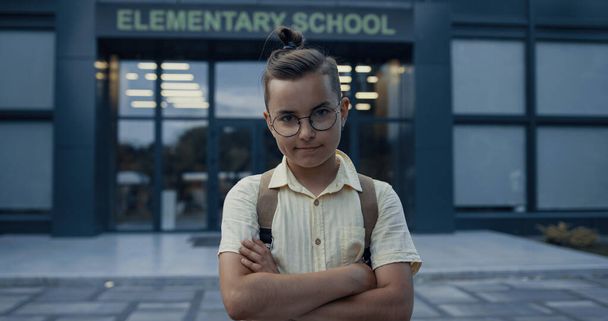 Cute elementary age guy posing on schoolyard alone close up. Serious little pupil with glasses standing at campus holding hands crossing. Portrait of smiling teen child wearing backpack looking camera - Photo, Image