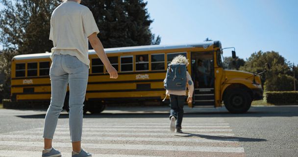 Радостная мама дает пять сыну сопровождения в школьный автобус. Молодая мать смотрит посадку на жёлтый автобус. Счастливый родитель сопровождает блондина в школу утром. Транспорт школьников, прибывающих на детском транспорте. - Фото, изображение