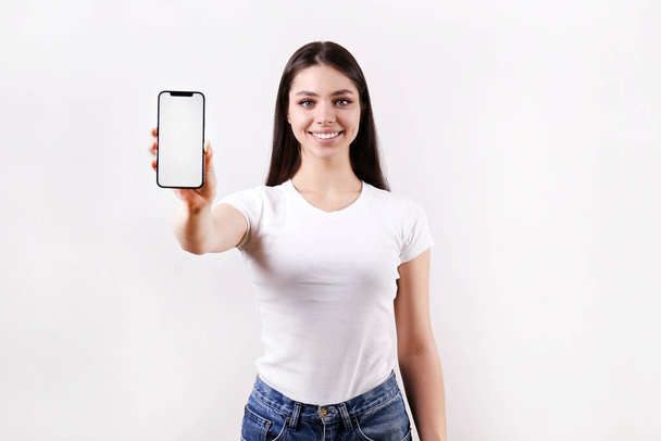 Young beautiful brunette woman with long hair wearing white t-shirt, posing with a phone over isolated background. Portrait of female model showing blank screen cellphone. Close up, copy space - Фото, изображение