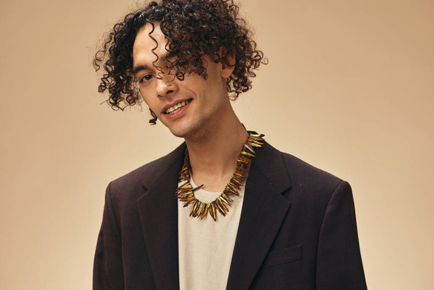 Flirting cute awesome tanned curly man in classic brown jacket ethnic necklace posing isolated on over beige pastel background. Fashion New Collection offer. Retro style concept. Free place for ad - 写真・画像