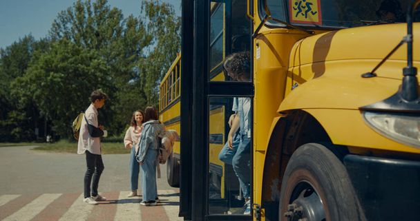 Two elementary school pupils leaving yellow classic bus chatting. Cheerful teenagers standing together talking near schoolbus. Teen classmates getting off academic shuttle. Education concept. - Photo, Image