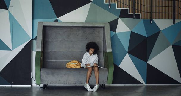 Preteen african american schoolgirl drawing in school corridor. Curly creative girl sitting bench writing poems in modern empty hall. Focused elementary age child creating sketches in notebook alone. - Fotoğraf, Görsel
