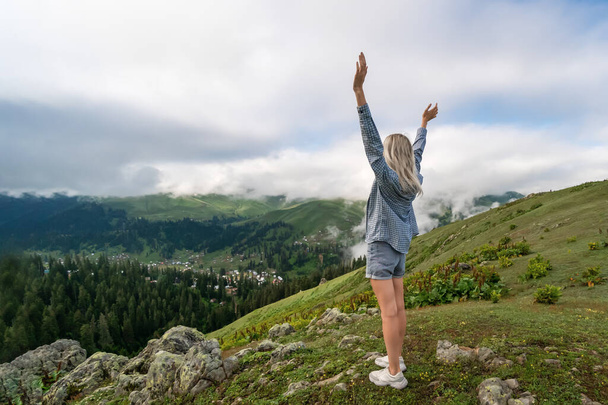 Young slender blonde woman in shorts and a shirt stands with her hands raised up on a mountain above the clouds. Bakhmaro, Georgia - Foto, Bild
