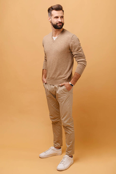 Full length of handsome calm brunette man wearing casual clothes looking at aside keeping hands in pockets isolated over beige background - Foto, imagen