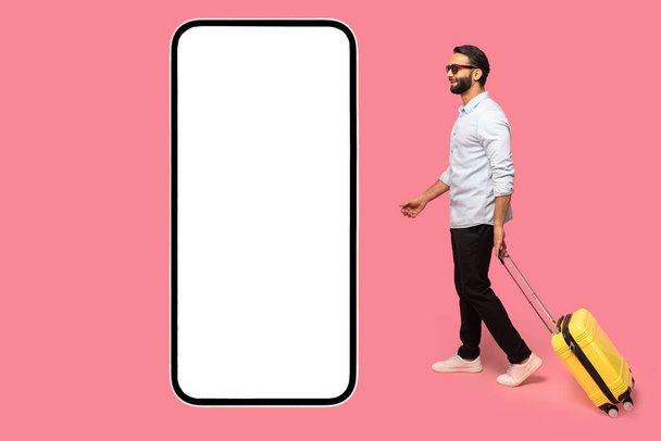 Mobile app for travelers. Indian man in sunglasses with yellow luggage suitcase walking to huge smartphone with empty display, copy space, mockup - Foto, immagini