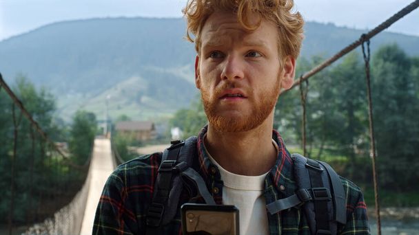 Sad traveler reading mobile phone screen. Hitchhiker looking map in mountains. Portrait of redhead man tourist exploring nature landscape background. Upset guy in inspiring journey. Holiday concept. - Photo, image