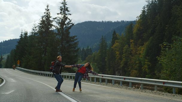 バックパッカーの山の道踊り。かわいいカップルは自然旅行の手を握っています。興奮した友人の森の高速道路で回っている。笑顔の旅行者は休日に一緒に時間を過ごす。レジャーコンセプト. - 写真・画像