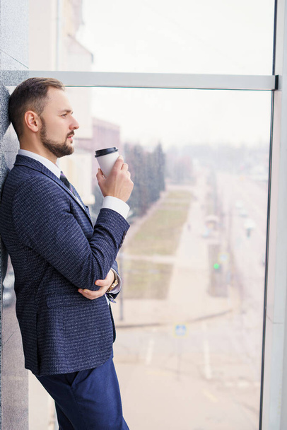 A young man with a neat little beard is dressed in a business suit with a white shirt and tie with a disposable paper cup of tea or coffee by the large window. Positive young businessman at the window - Foto, Bild