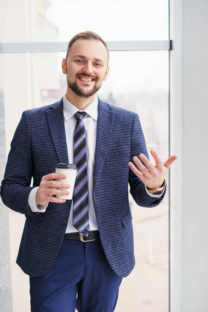 A young man with a neat little beard is dressed in a business suit with a white shirt and tie with a disposable paper cup of tea or coffee by the large window. Positive young businessman at the window - Photo, Image