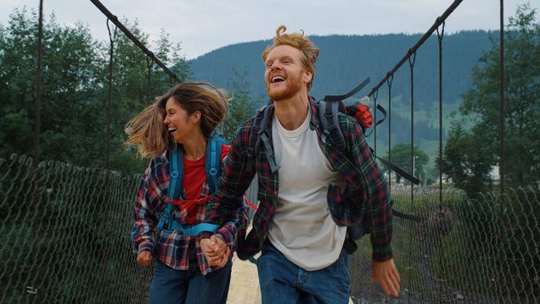 感情的なカップル旅行者の山の橋の上を実行している。笑顔のハイカーは外で速くレースします。野生の自然の中で自由を感じて興奮した観光客。旅行で手をつないで楽しい愛好家。休日のコンセプト.  - 写真・画像