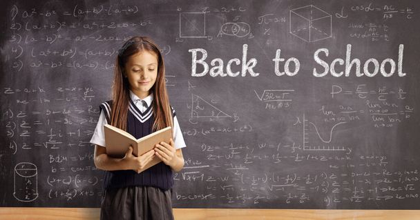 Schoolgirl reading a book in front of a blackboard with math formulas and text back to school - Photo, image