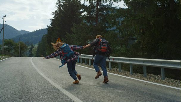 山の道で森の自然を踊る恋人たち。二人のヒッチハイカーは外で手を握っている。正のバックパッカーの空の高速道路でジャンプします。ハイキングで幸せを感じる笑顔のヒップスター。幸福の概念. - 写真・画像