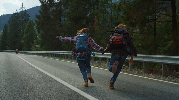 Couple run mountains countryside on roadside. Smiling hikers hold hands in forest nature. Happy lovers celebrate marriage honeymoon outdoors. Travelers dancing on highway. Having fun leisure concept. - Photo, Image