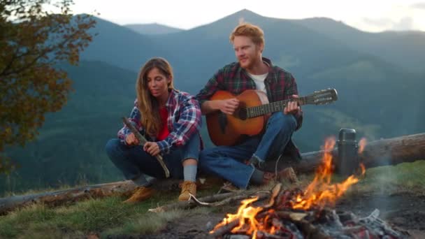 Relaxed tourists enjoy bonfire spend evening in mountains. Close up happy couple camp outdoors on sunset nature. Romantic lovers play guitar on holiday weekend. Family activity together concept. - Filmati, video