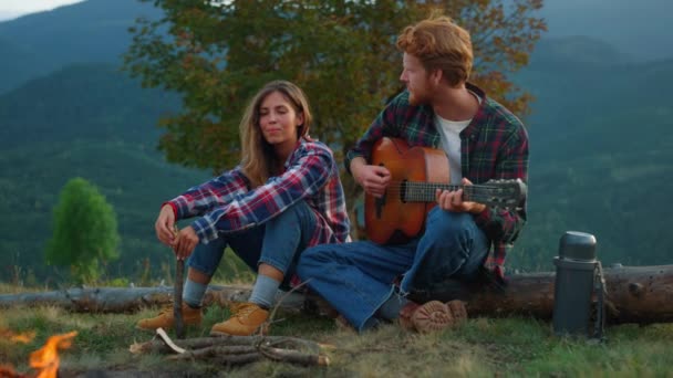 Closeup couple travelers relax together on mountains trip. Lovely campers enjoy romantic music. Affectionate lovers look each other on evening bonfire. Chill tourists play guitar. Leisure concept. - Video, Çekim