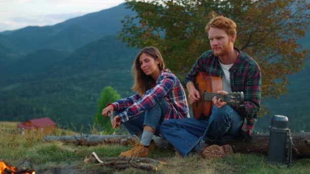 Positive couple date outdoors in mountains. Close up two lovers play guitar by campfire. Carefree travelers relax on activity. Resting campers enjoy music on romantic evening picnic. Relax concept. - Filmati, video