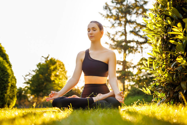 Young peaceful woman sitting in lotus pose, breathing and meditating. Concept of outdoors yoga, relaxation, zen and mental balance. Beautiful yogini in black sportswear in the park at sunset. - Photo, image