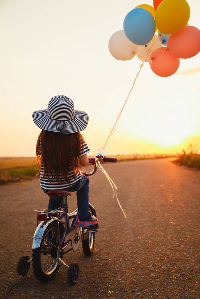 Cute little girl with colorful balloons cycling at the road at sunset sky summer time. Back view - Foto, afbeelding