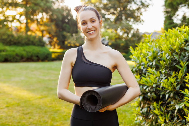 Portrait of young smiling woman holding in hands black sports mat. Concept of outdoors yoga, fitness, active and sporty lifestyle - Foto, afbeelding