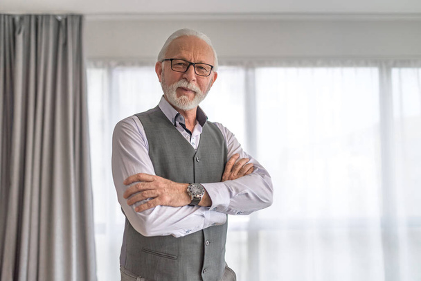Portrait of confident senior businessman. Elderly male professional is standing with arms crossed against window. He is wearing formals at corporate office. - Photo, image