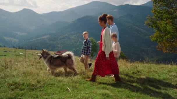 Счастливые родители с детьми выгуливают дружелюбную собаку на горном холме солнечным днем. Симпатичная девушка с матерью, держащей поводок на зеленой траве. Расслабленная семья с детьми, отдыхающими на свежем воздухе - Кадры, видео