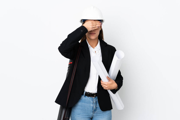 Young architect caucasian woman with helmet and holding blueprints over isolated background covering eyes by hands - Photo, Image