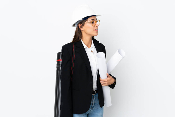 Young architect caucasian woman with helmet and holding blueprints over isolated background looking to the side - Foto, Imagen