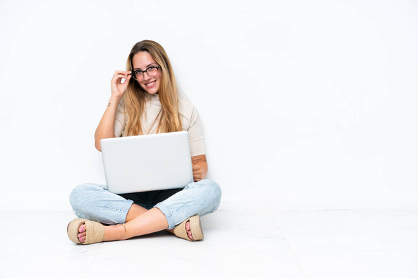 Jovem mulher com laptop sentado no chão isolado no fundo branco com óculos e feliz - Foto, Imagem