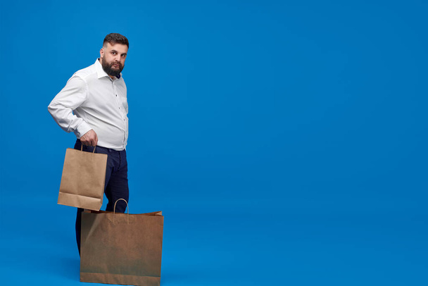A Caucasian-looking man with a brown paper bag on a blue background. A man with a white shirt with shopping bags. Studio shot of a man with purchases. - Fotó, kép