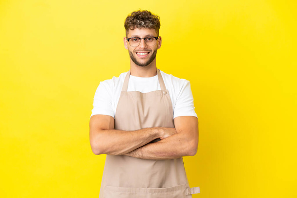 Restaurant waiter blonde man isolated on yellow background keeping the arms crossed in frontal position - Foto, Imagen