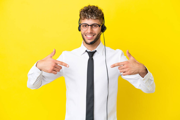 Telemarketer blonde man working with a headset isolated on yellow background proud and self-satisfied - Foto, afbeelding