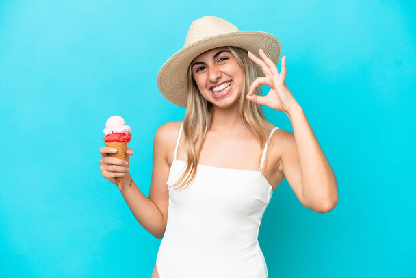 Young caucasian woman in swimsuit with a cornet ice cream isolated on blue background showing ok sign with fingers - Fotoğraf, Görsel