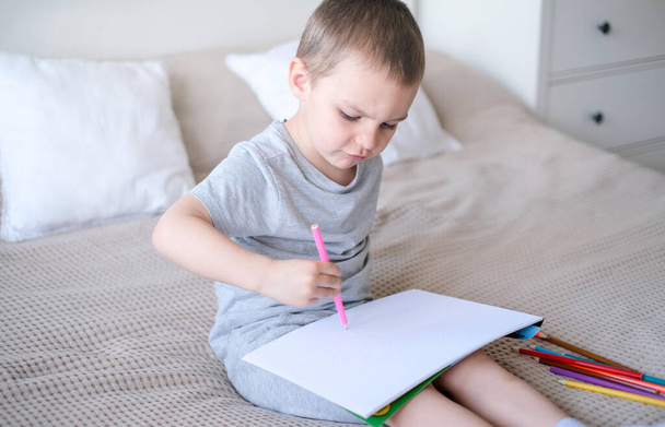 A small caucasian boy in a gray T-shirt and shorts on the bed draws with colored pencils in a sketchbook. Childrens creativity and development. - Foto, afbeelding