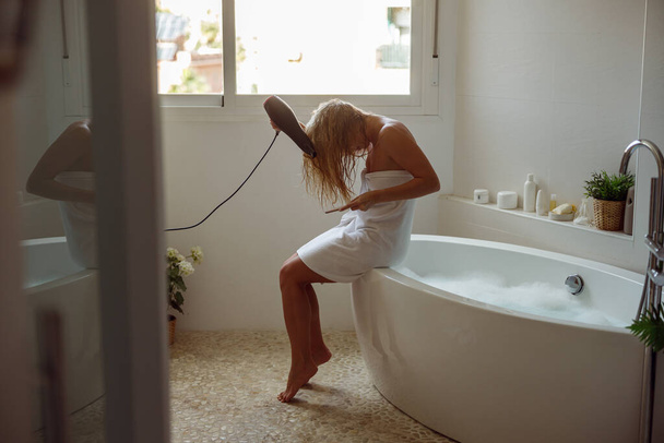 Pretty Caucasian female drying hair with dryer after taking bath. Morning beauty routine. Bathroom. - Zdjęcie, obraz
