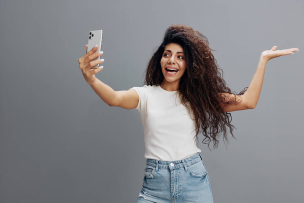 Boldog göndör nő okostelefon, hogy videohívás, vagy hogy Selfie, portré vidám afro-amerikai nő lyukas kézzel a mobiltelefon kamera, miközben áll át Gray Studio háttér - Fotó, kép