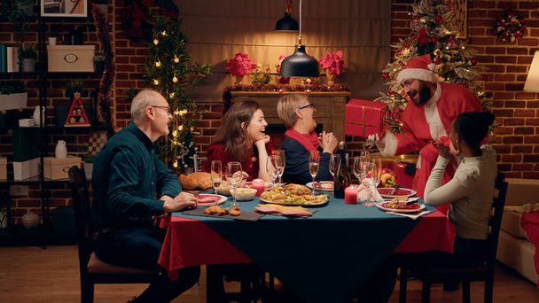 Wholesome man disguised as Santa Claus giving festive family presents at Christmas dinner. Funny husband wearing Santa Claus costume offering gifts to family while celebrating winter holiday at home. - Fotografie, Obrázek