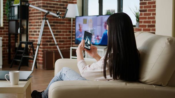Childish young adult person enjoying mobile gaming while sitting on sofa inside apartment. Playful asian woman playing space shooter game on smartphone device while working remotely from home. - Photo, Image
