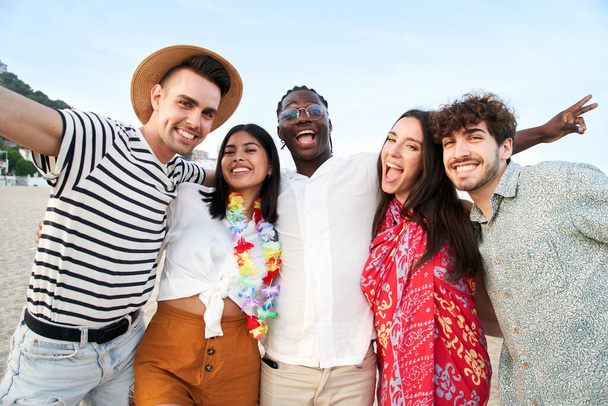 Portrait of five young multicultural friends on the sea shore looking at camera laughing. Multiracial young women and men strolling along a beach. High quality photo - Foto, Imagem