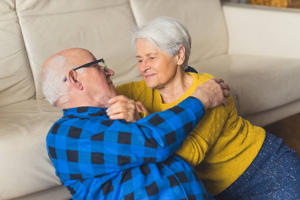 Relaxed beautiful loving embracing senior family couple looking at each other and smiling while sitting on the floor near the couch in the living room at home. High quality photo - Photo, image