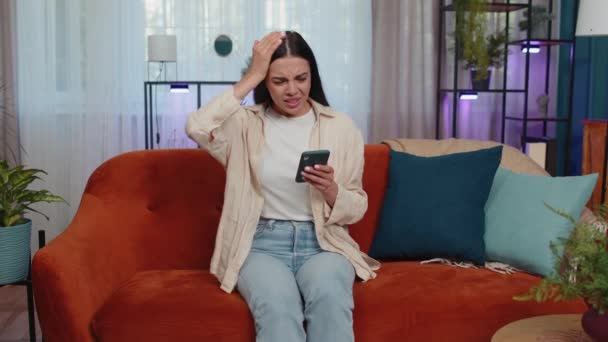 Portrait of caucasian adult girl use smartphone typing browsing, loses becoming surprised sudden lottery results, bad news, fortune loss, fail. Young woman sitting on orange couch at home living room - Materiaali, video