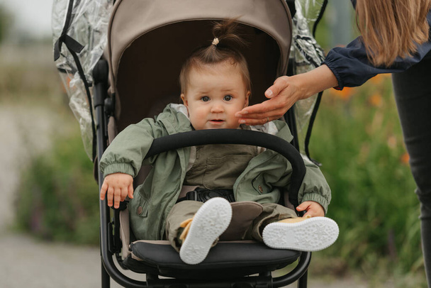 A female toddler is sitting in the stroller on a cloudy day. In a green village, a young serious girl in a raincoat is in a baby carriage. A mother is touching the cheek of her daughter. - Photo, Image