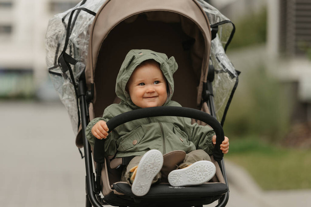 A female toddler is holding the safety bumper bar of the stroller on a cloudy day. A young girl in the hood of her raincoat is in her baby carriage in a park at noon. - Foto, Imagen