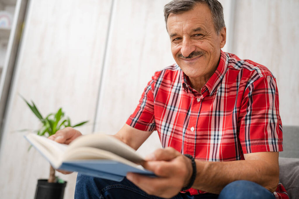 Ένας άνδρας ανώτερος καυκάσιος άνδρας με μουστάκια διαβάζει βιβλίο στο σπίτι φορά κόκκινο πουκάμισο πραγματικοί άνθρωποι έννοια αναψυχής αντίγραφο χώρο κοιτάζοντας προς την κάμερα χαρούμενο χαμόγελο - Φωτογραφία, εικόνα