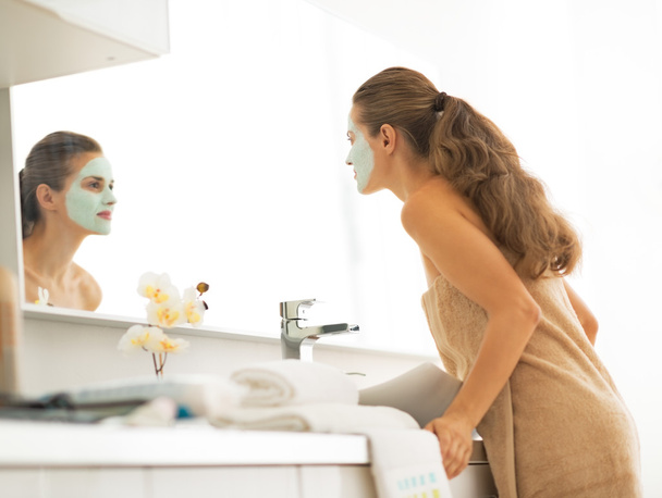 Mujer joven con máscara cosmética facial mirando en el espejo en bathr
 - Foto, Imagen