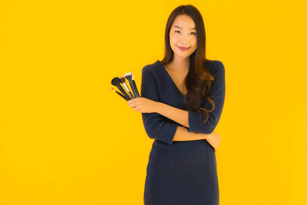 Porträt schöne junge asiatische Frau mit Make-up-Pinsel auf gelbem Hintergrund - Foto, Bild