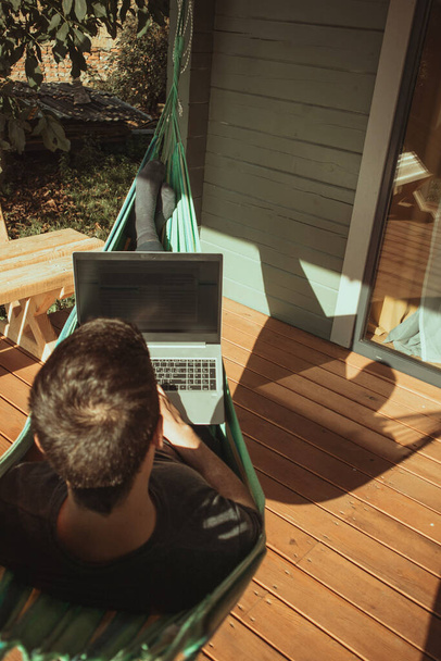 Een man freelancer ligt in een hangmat en werkt in de buurt van het landhuis op een zonnige dag. Afstandsbediening - Foto, afbeelding