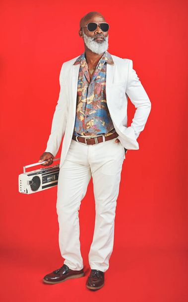 People hear me coming before they see me. Studio shot of a senior man wearing vintage clothing while posing with a boombox against a red background - Foto, Imagen